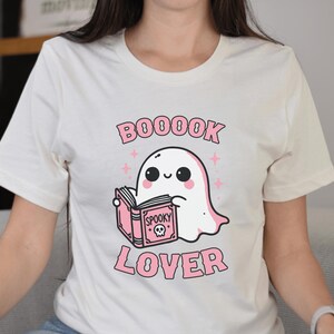 May include: Cream-colored t-shirt featuring a cute cartoon ghost reading a pink book with the words "Spooky" and a skull on the cover. The shirt also has the words "BOOOOK LOVER" in pink.
