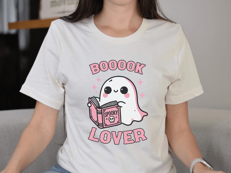 May include: Cream-colored t-shirt featuring a cute cartoon ghost reading a pink book with the words "Spooky" and a skull on the cover. The shirt also has the words "BOOOOK LOVER" in pink.