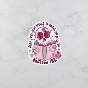 Könnte beinhalten: Ein Aufkleber mit einem rosa und lila Schädel, der ein Buch liest, mit dem Text "Shhh, I'm just trying to catch up with my Endless TBR." Der Schädel ist von Blumen umgeben. Das Buch ist rosa.