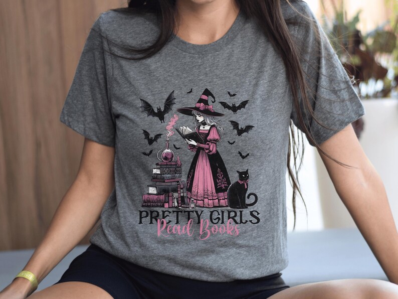 May include: Gray t-shirt featuring a witch reading a book, surrounded by bats and a black cat. The witch wears a black hat and pink dress. The text "PRETTY GIRLS Read Books" is printed below the illustration.
