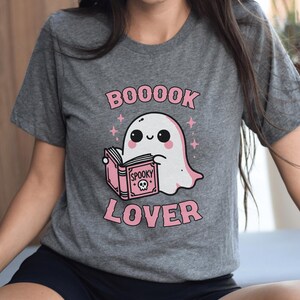 May include: Gray t-shirt featuring a cute ghost reading a pink book with the words "Spooky" and a skull on the cover. The shirt has the words "BOOOOK LOVER" in pink above and below the ghost illustration.