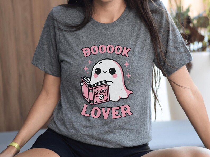 May include: Gray t-shirt featuring a cute ghost reading a pink book with the words "Spooky" and a skull on the cover. The shirt has the words "BOOOOK LOVER" in pink above and below the ghost illustration.