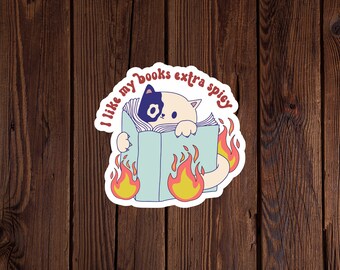 May include: A white sticker with a black and white cat reading a book with flames coming out of the book. The text on the sticker reads "I like my books extra spicy".