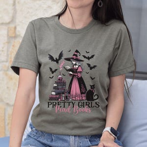 May include: Olive green t-shirt featuring a witch reading a book, surrounded by bats, a black cat, and a stack of books. The text "PRETTY GIRLS Read Books" is printed below the illustration. The witch is wearing a pink and black dress.