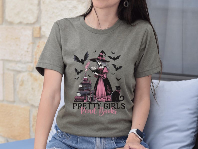 May include: Olive green t-shirt featuring a witch reading a book, surrounded by bats, a black cat, and a stack of books. The text "PRETTY GIRLS Read Books" is printed below the illustration. The witch is wearing a pink and black dress.