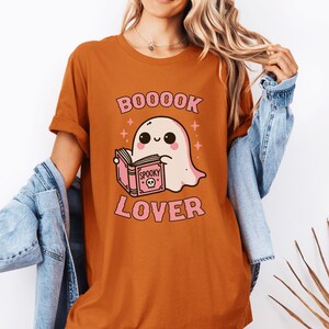 May include: Orange t-shirt with a cartoon ghost reading a book. The shirt has the text "BOOOOK LOVER" in pink, with a cute ghost illustration and the word "Spooky" on the book. The shirt is a Halloween-themed design.