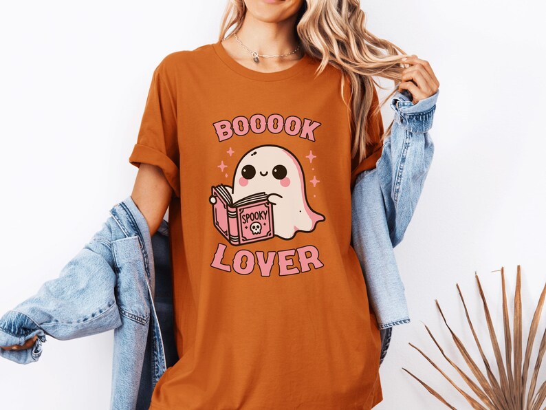 May include: Orange t-shirt with a cartoon ghost reading a book. The shirt has the text "BOOOOK LOVER" in pink, with a cute ghost illustration and the word "Spooky" on the book. The shirt is a Halloween-themed design.