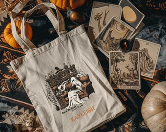 Puede incluir: Bolso tote beige con un fantasma leyendo en un sofá, ambientado en una biblioteca. El bolso tiene el texto "Born To Read" y "KAYLEIGH". Calabazas, cartas del tarot y una vela completan la escena de Halloween.