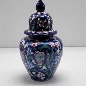 May include: A dark blue ceramic jar with a lid, adorned with a floral pattern in red, white, and blue. The jar has a bulbous body and a lid with a pointed finial. The floral design includes leaves and small flowers.