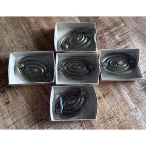 May include: Six antique brass oval drawer pulls in individual white cardboard boxes. Each pull has a decorative center and two screw holes. The pulls are displayed on a wooden surface.