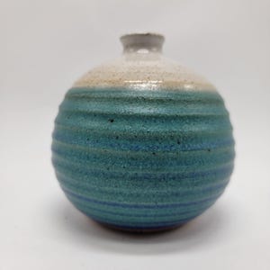 May include: A spherical ceramic vase with a narrow neck, featuring a textured, ribbed surface. The glaze transitions from a creamy off-white at the top to a deep, speckled teal and blue gradient across the rounded body of the vessel.