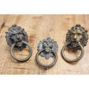May include: Three antique bronze lion head door knockers with ring handles. The lion heads have detailed manes and open mouths. The knockers are arranged on a wooden surface.