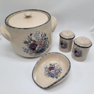 May include: A cream-colored ceramic serving set featuring a large lidded pot with handles, two salt and pepper shakers, and a small serving dish. Each piece is decorated with a floral design in shades of purple, blue, and pink, with a blue trim.