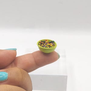 May include: A miniature lime green bowl filled with colorful, tiny, speckled pieces. The bowl is held between two fingers against a white background. The bowl's contents appear to be a mix of various colors.