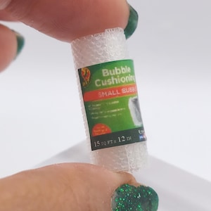 May include: A miniature roll of clear bubble cushioning is held between fingers. The label reads "Bubble Cushioning" and "SMALL BUBBLE" with the dimensions "15 sq ft x 12 in". The roll is a crafting or packaging supply.