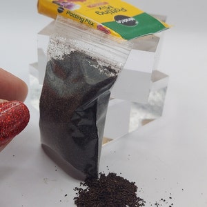 Pode incluir: Um saco pl&aacute;stico transparente cheio de sementes castanhas escuras, com uma pequena pilha de sementes derramadas. Um pacote de mistura para vasos amarelo e verde ao fundo. A imagem est&aacute; em uma superf&iacute;cie branca.