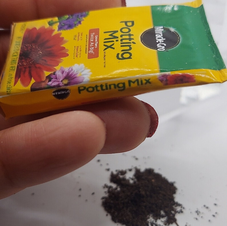 Pode incluir: Um pequeno pacote de mistura para vasos Miracle-Gro, com um design amarelo e verde. O pacote apresenta imagens de flores coloridas e o texto "Twice As Big!". Uma pequena pilha de mistura para vasos &eacute; vis&iacute;vel abaixo.