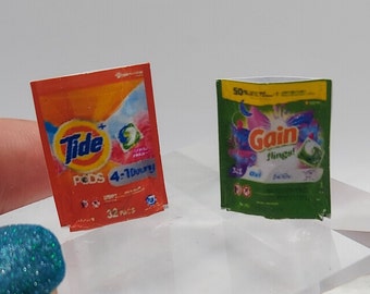 May include: Two miniature laundry detergent boxes. The Tide box is orange and features a blue and white logo with the text "Tide" and "PODS 4-in-1" in white. The Gain box is green and features a white and blue logo with the text "Gain flings!" in white.