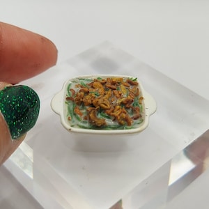 May include: Miniature green bean casserole in a white dish with handles. The casserole has a creamy sauce, green beans, and crispy fried onions on top. A tiny, detailed food item.