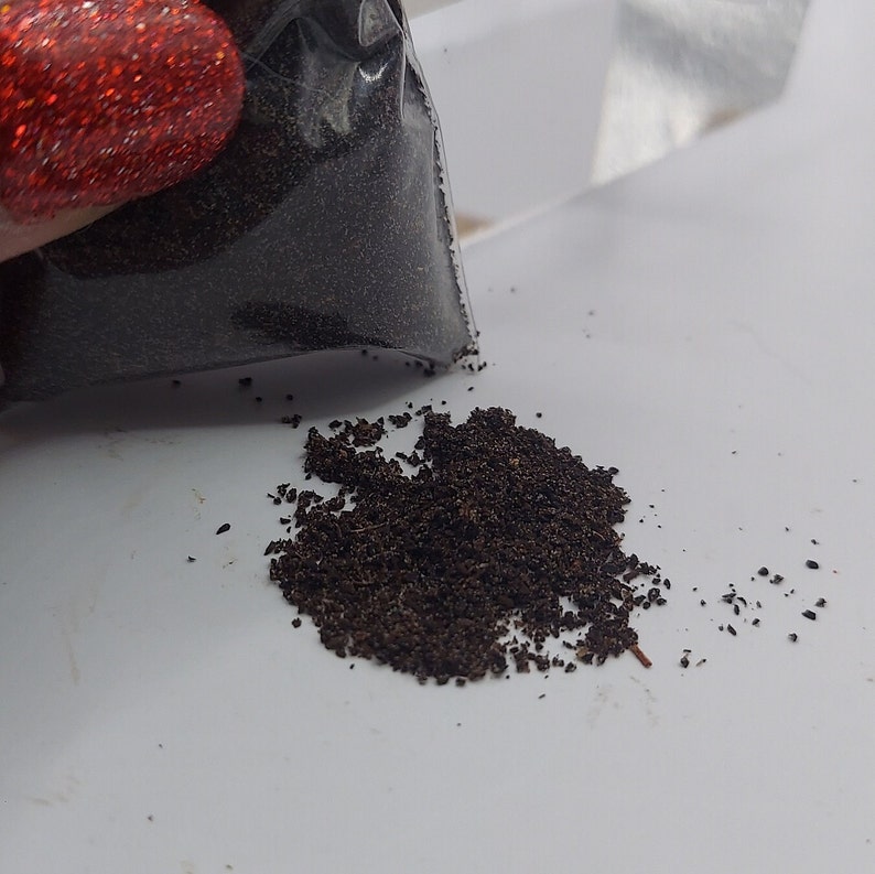 Pode incluir: Um saco pl&aacute;stico transparente parcialmente preenchido com gr&acirc;nulos castanhos escuros, com alguns gr&acirc;nulos derramados sobre uma superf&iacute;cie branca. Os gr&acirc;nulos s&atilde;o pequenos e parecem ser um p&oacute; fino ou material mo&iacute;do.