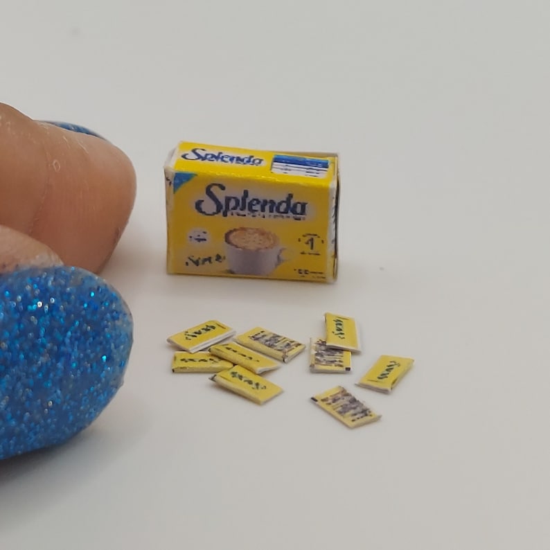 May include: A miniature yellow box and packets of Splenda sweetener. The box has the Splenda logo and an image of a cup of coffee. Several small yellow packets are scattered around the box.