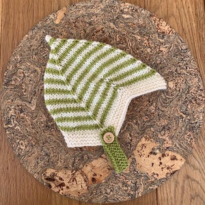 May include: A pointed knit bonnet featuring horizontal stripes in light green and off-white. The bonnet has a garter stitch border along the face opening and a single green knit chin strap secured with a round, light brown wooden button.