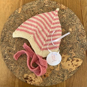May include: A hand-knitted bonnet featuring a pink and cream striped pattern with a pointed top. The bonnet includes matching pink knitted chin ties and a white circular tag attached with a white ribbon, displaying the text Alpaca and a graphic.