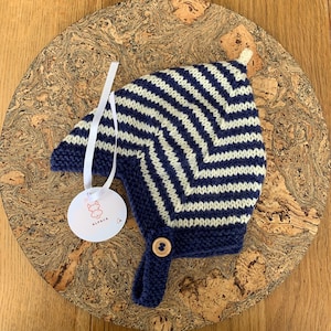 May include: A knitted bonnet featuring a navy blue and cream chevron pattern with a solid navy blue border. The bonnet includes a chin strap secured by a light brown wooden button and a white circular tag with the text ALPACA and a line drawing.