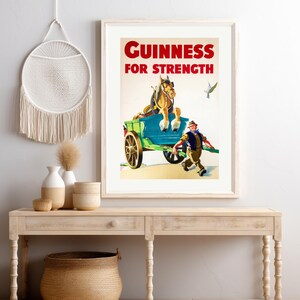 K&ouml;nnte beinhalten: Gerahmtes Vintage-Guinness-Werbeposter. Das Poster zeigt einen Pferdewagen mit dem Text "Guinness For Strength" in Rot. Ein Mann rennt neben dem Wagen, und ein Vogel fliegt im Hintergrund. Das Poster wird auf einem hellen Holztisch pr&auml;sentiert.