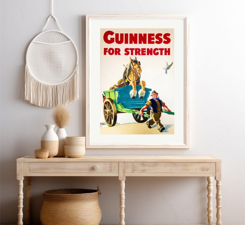 K&ouml;nnte beinhalten: Gerahmtes Vintage-Guinness-Werbeposter. Das Poster zeigt einen Pferdewagen mit dem Text "Guinness For Strength" in Rot. Ein Mann rennt neben dem Wagen, und ein Vogel fliegt im Hintergrund. Das Poster wird auf einem hellen Holztisch pr&auml;sentiert.