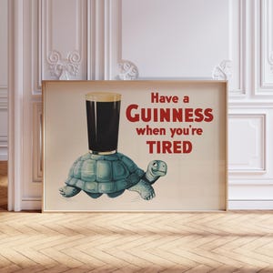 K&ouml;nnte beinhalten: Ein gerahmter Druck mit einem Guinness-Pint auf einer t&uuml;rkisfarbenen Schildkr&ouml;te. Der rote Text lautet "Have a Guinness when you're TIRED". Das Bild hat einen Vintage-Cartoon-Stil.