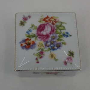 May include: A square white porcelain trinket box featuring a colorful floral bouquet on the lid. The design includes pink roses, blue forget-me-nots, and orange blossoms. The edges of the lid are finished with a delicate gold trim.