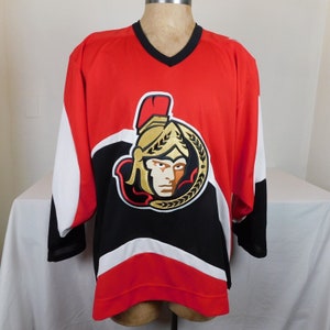 May include: A red, white, and black hockey jersey with the Ottawa Senators logo. The logo features a gold and black gladiator helmet with a red plume.