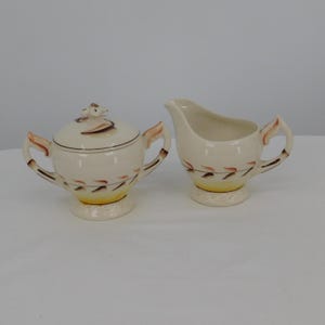 May include: Cream-colored ceramic sugar bowl with lid and creamer set. The sugar bowl has a lid with a decorative handle. Both pieces feature brown and orange leaf-like designs and sit on a textured base. The creamer has a curved spout.