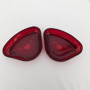 May include: Two red, teardrop-shaped automotive taillight lenses. Each lens has a black border and a layered, geometric design. The lenses are made of a translucent material, allowing light to pass through.