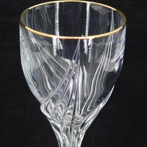 May include: A clear wine glass with a gold rim. The glass features a detailed, swirling design on the bowl. The stem is slender and elegant. The glass is set against a black background.