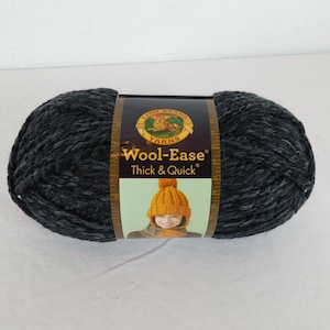 Op de afbeelding: Een bol donker antracietgrijs Lion Brand Wool-Ease Thick & Quick garen met een centraal papieren label. Het label bevat het merklogo, de productnaam en een illustratie van een persoon die een mosterdgele gebreide muts draagt.