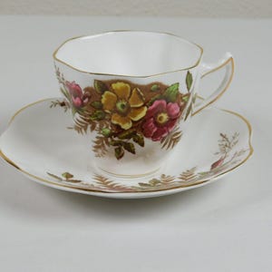 May include: A white teacup and saucer with a floral pattern. The pattern features pink, yellow, and brown flowers with green leaves on a white background. The cup and saucer have a gold trim.
