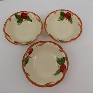 Puede incluir: Tres cuencos de cerámica blanquecina con borde festoneado. Cada cuenco presenta un diseño pintado de una fruta roja con hojas verdes y un borde marrón en forma de rama. Los cuencos están dispuestos sobre una superficie blanca.