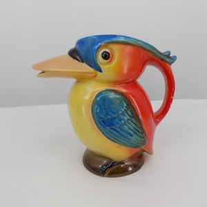 May include: A colorful ceramic pitcher shaped like a bird. The pitcher features a yellow body, a blue head, and red accents. The bird has a long, tan beak and a curved handle. The base is brown.