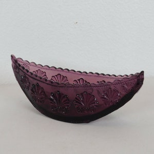 May include: A deep purple, oval-shaped glass bowl. The bowl features a scalloped rim and is decorated with embossed seashell designs. The glass has a glossy finish and is set against a white background.