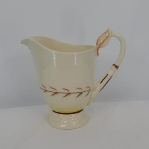 May include: Cream-colored ceramic pitcher with a leaf-like handle and a decorative leaf pattern around the body. The pitcher has a flared spout and a pedestal base. The handle is brown and the leaf pattern is brown and orange.