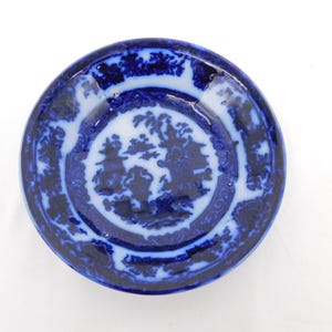 May include: A round, antique blue and white plate with a detailed landscape design in the center. The plate has a dark blue border with floral and scroll patterns. The central design features buildings and trees in a classic style.