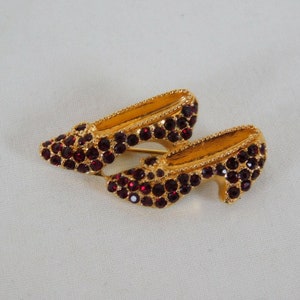 Peut inclure: Une broche dorée en forme de deux chaussures à talons hauts. Les chaussures sont recouvertes de strass rouges.