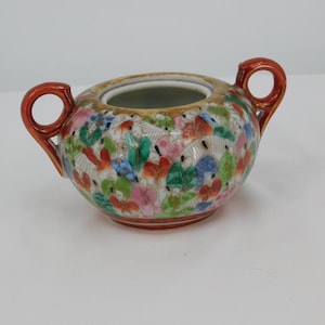 Puede incluir: Un pequeño azucarero de porcelana blanca con tapa y dos asas rojas. El bol está decorado con un patrón floral colorido y detalles dorados.