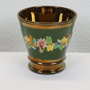 May include: A small, copper-coloured ceramic vase with a green band and a floral design. The vase has a flared rim and a rounded base.