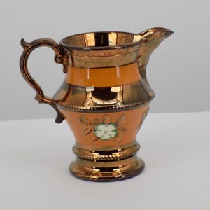 Puede incluir: Una jarra de cerámica antigua con asa y boquilla color bronce. La jarra presenta un cuerpo naranja con bandas de bronce y un diseño floral. La base y el borde también son de color bronce.