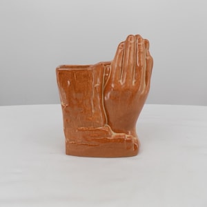 May include: A glossy, light brown ceramic planter shaped like two hands pressed together in a prayer position. The surface features a smooth, reflective glaze with subtle variations in color tone, and the top is open to hold small items or plants.