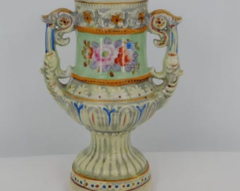 May include: A decorative ceramic vase with a light green central panel featuring a floral design in pink, red, and blue. The vase has two ornate handles and a pedestal base. The vase is approximately 8 inches tall.