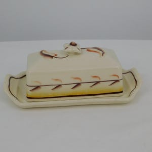May include: Cream-colored ceramic butter dish with a rectangular lid and a matching tray. The lid features brown and orange leaf-like designs and a small flower-shaped handle. The tray and lid are trimmed with a brown border.